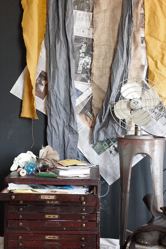 Still life with antique flat file cabinet, old electric fan, and layered textiles against a dark wall in an indoor setting.