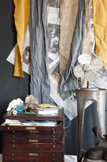Still life with antique flat file cabinet, old electric fan, and layered textiles against a dark wall in an indoor setting.