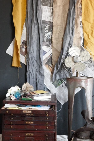 Still life with antique flat file cabinet, old electric fan, and layered textiles against a dark wall in an indoor setting.
