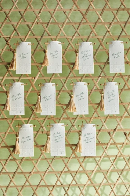 Wedding seating chart on a wooden lattice with small cards featuring guest names and gold tassels. Elegant reception decor.