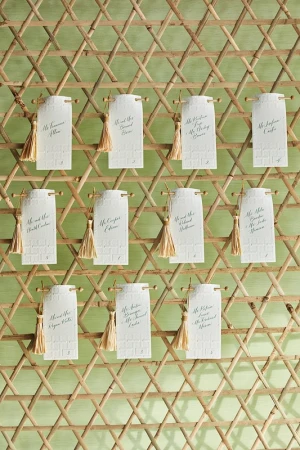Wedding seating chart on a wooden lattice with small cards featuring guest names and gold tassels. Elegant reception decor.