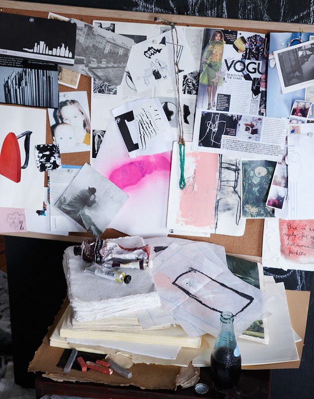 Artist's workspace featuring a corkboard with pinned artwork and magazine covers, set atop a desk with art supplies and a vintage soda bottle.