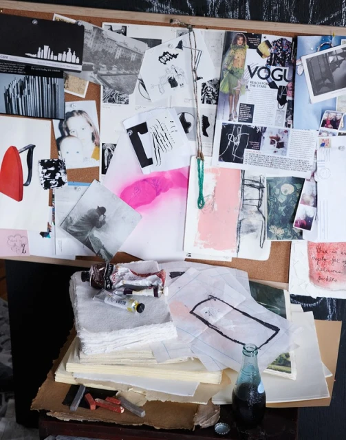Artist's workspace featuring a corkboard with pinned artwork and magazine covers, set atop a desk with art supplies and a vintage soda bottle.