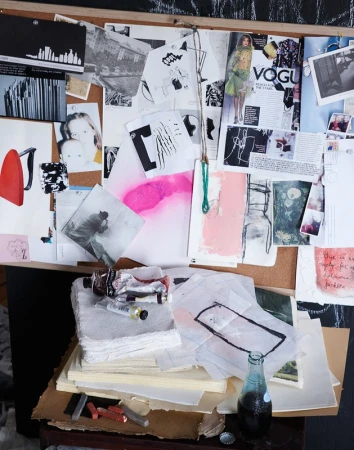 Artist's workspace featuring a corkboard with pinned artwork and magazine covers, set atop a desk with art supplies and a vintage soda bottle.