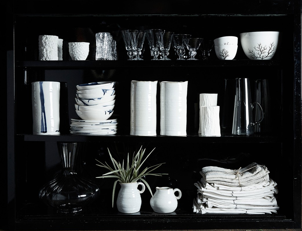 Display cabinet showcasing an arrangement of white ceramic vessels, glassware, bowls, and folded textiles in a dark, modern setting.