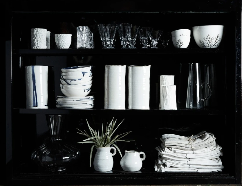 Display cabinet showcasing an arrangement of white ceramic vessels, glassware, bowls, and folded textiles in a dark, modern setting.