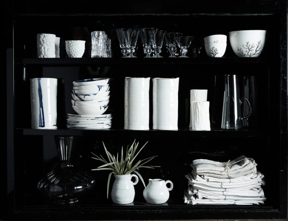 Display cabinet showcasing an arrangement of white ceramic vessels, glassware, bowls, and folded textiles in a dark, modern setting.