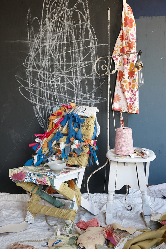 Eclectic composition featuring painted stools, assorted fabrics, and an apron against a dark wall with white scribbles, creating a whimsical.