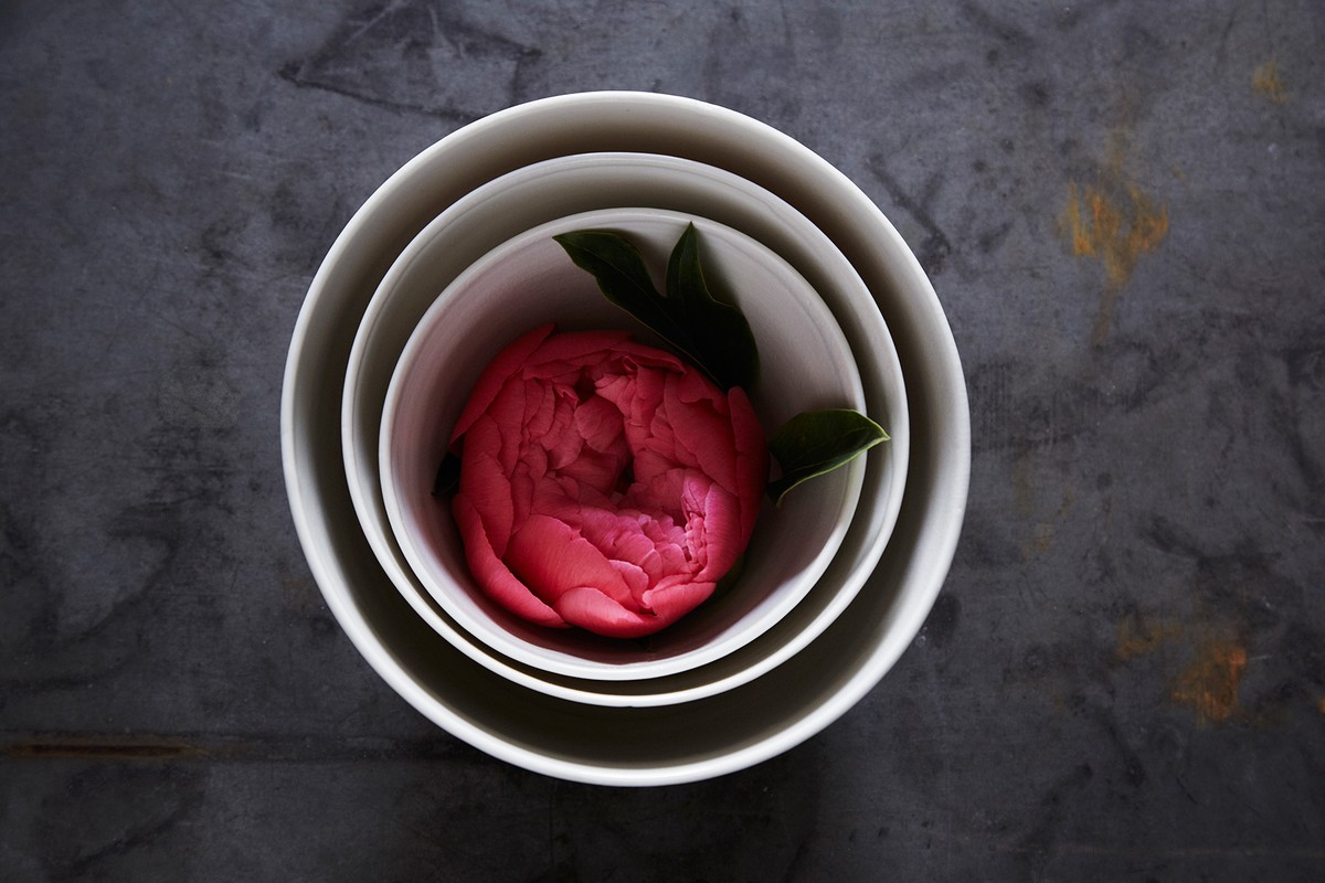 Overhead shot of a pink peony flower with green leaves centered in a set of three graduated white ceramic bowls on a dark gray surface.