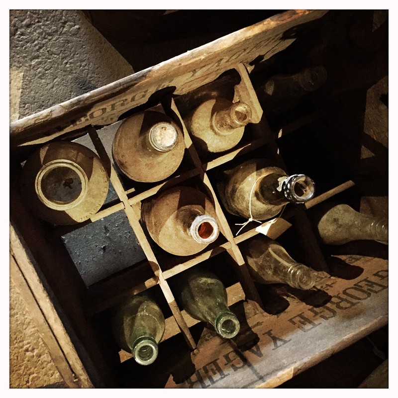 Still life photo of dusty empty glass bottles in a wooden crate with dividers in a dimly lit, aged cellar. Focus on vintage storage.