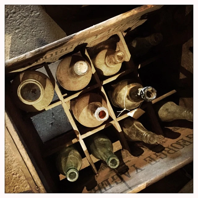 Still life photo of dusty empty glass bottles in a wooden crate with dividers in a dimly lit, aged cellar. Focus on vintage storage.