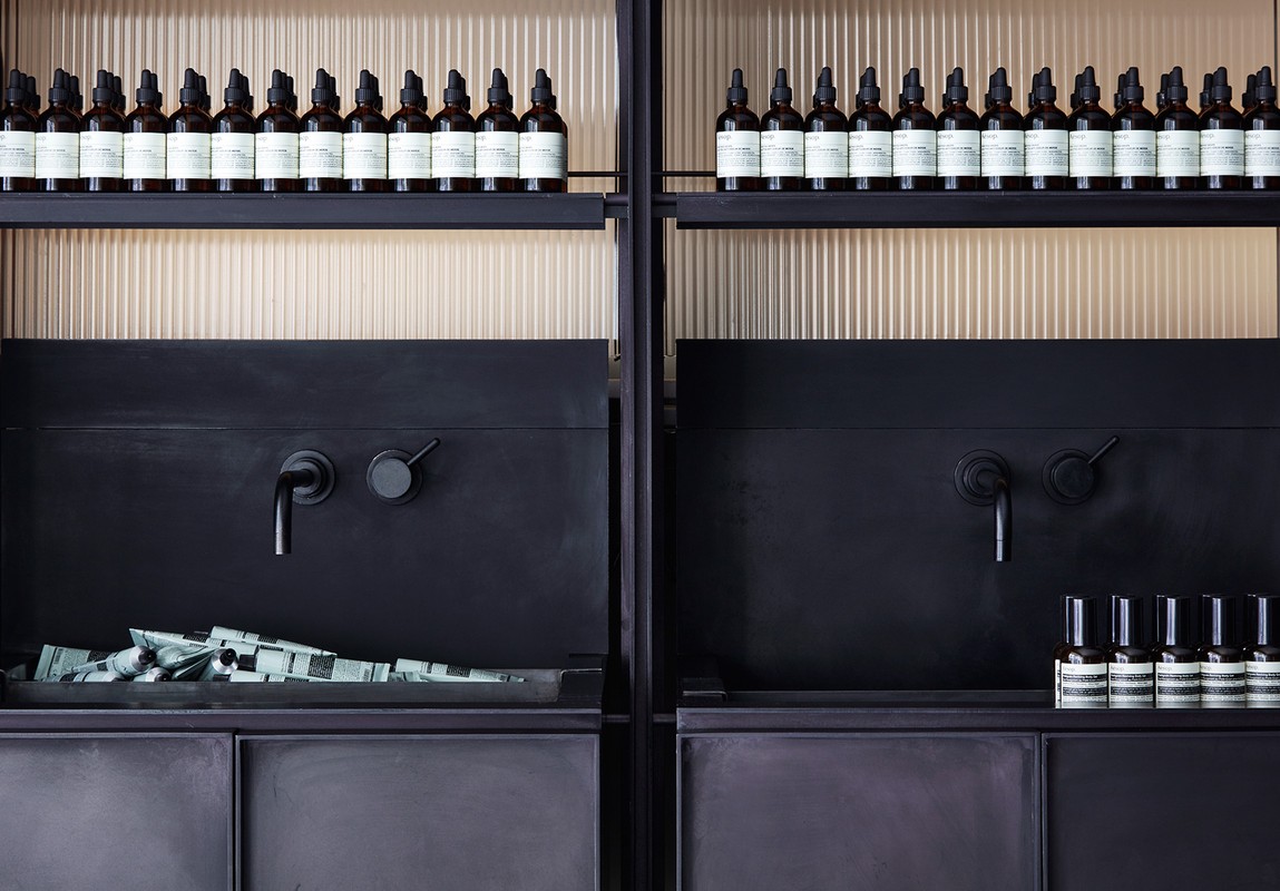 Dark interior with symmetrically arranged beauty products on shelves: bottles with droppers, tubes, and spray bottles against a vertically textured wall.