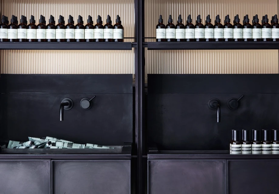 Dark interior with symmetrically arranged beauty products on shelves: bottles with droppers, tubes, and spray bottles against a vertically textured wall.