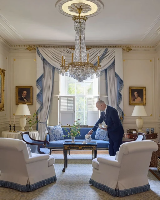 Elegant interior featuring an adult male in a suit serving tea or coffee on a blue sofa beneath a crystal chandelier, bathed in natural light.
