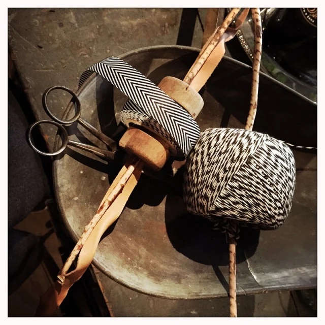 Still life of a spool of black and white thread, scissors, wooden dowel, and chevron ribbon on a tarnished metal surface, vintage aesthetic.