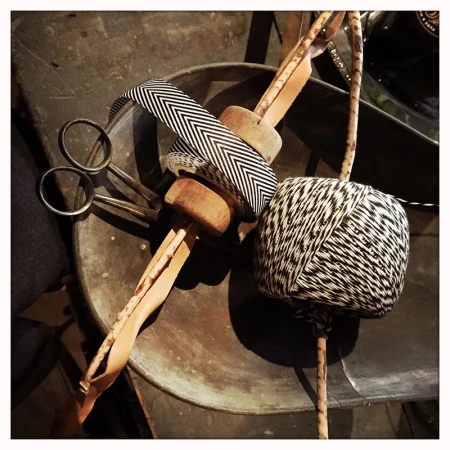 Still life of a spool of black and white thread, scissors, wooden dowel, and chevron ribbon on a tarnished metal surface, vintage aesthetic.