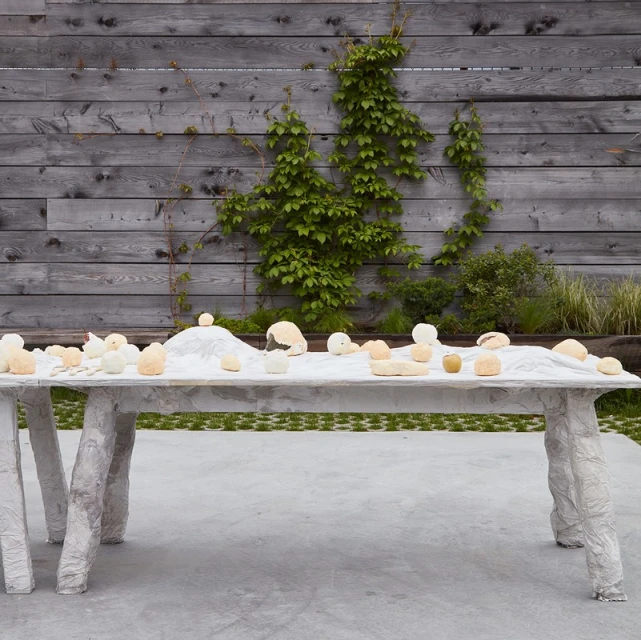 Outdoor still life features white tablecloth-covered table with white, tan, and apple-green objects and a rustic wood fence backdrop with vines.