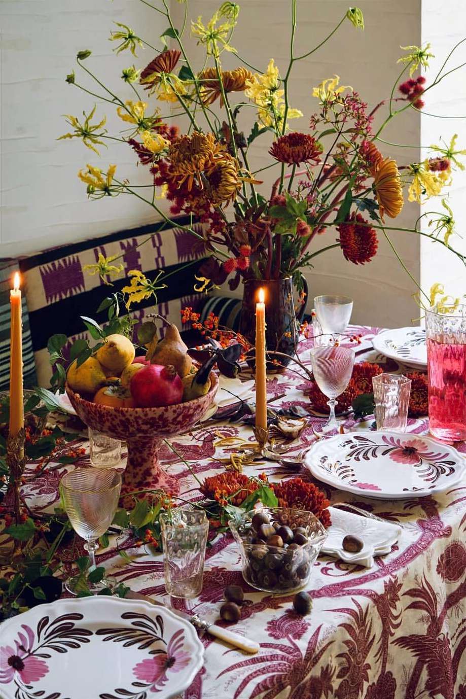 Table setting with floral centerpiece, fruit bowl, candles, and patterned tablecloth creating a warm, inviting ambiance.