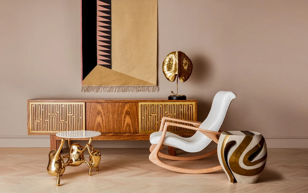 Living room interior featuring wood cabinet, side tables with metallic accents, rocking chair, gold leaf sculpture, and geometric wall hanging.