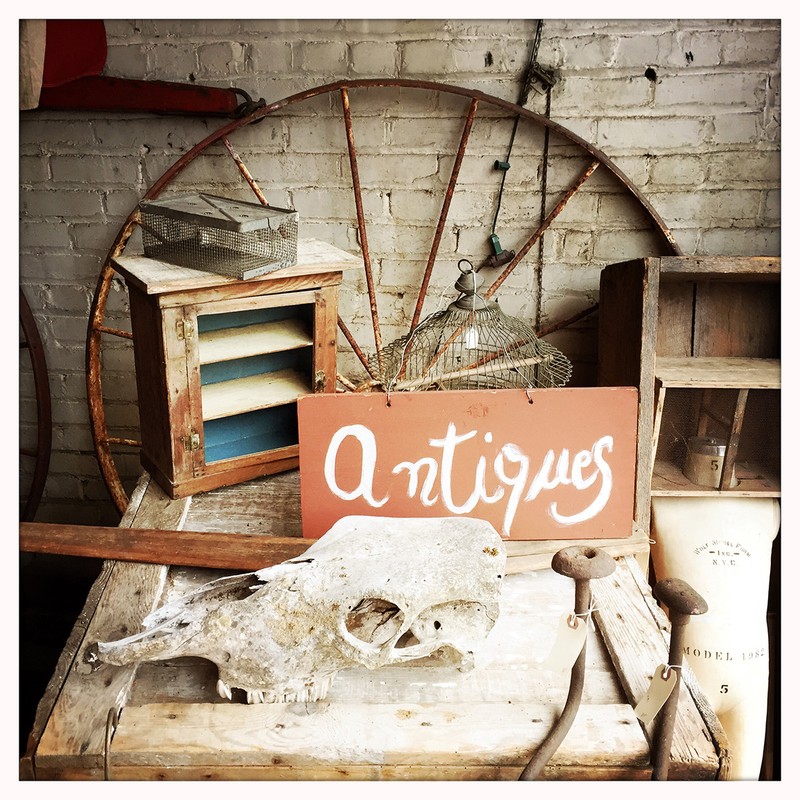 Rustic antique display features an old sign, animal skull, vintage cabinet, and metal wheel against a brick wall indoors.