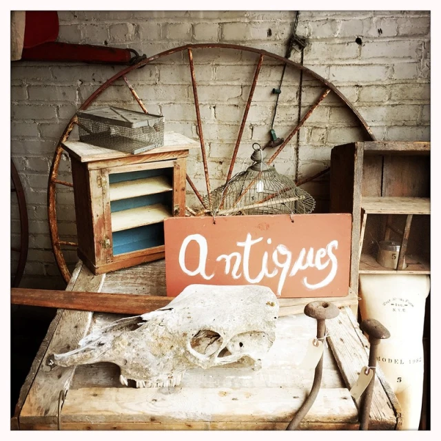 Rustic antique display features an old sign, animal skull, vintage cabinet, and metal wheel against a brick wall indoors.