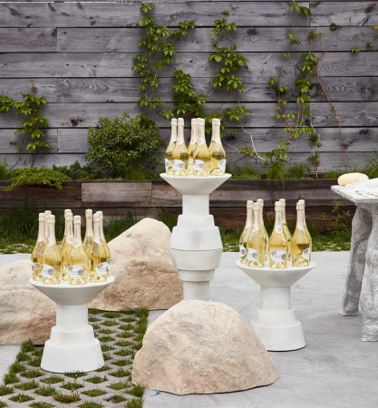 Champagne bottles displayed on pedestals in an outdoor setting against a rustic wooden fence with vines. Decorative stone elements.