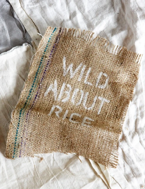 Burlap sack fragment with white lettering 'Wild About Rice' displayed on rumpled white fabric, top-down angle, natural light.