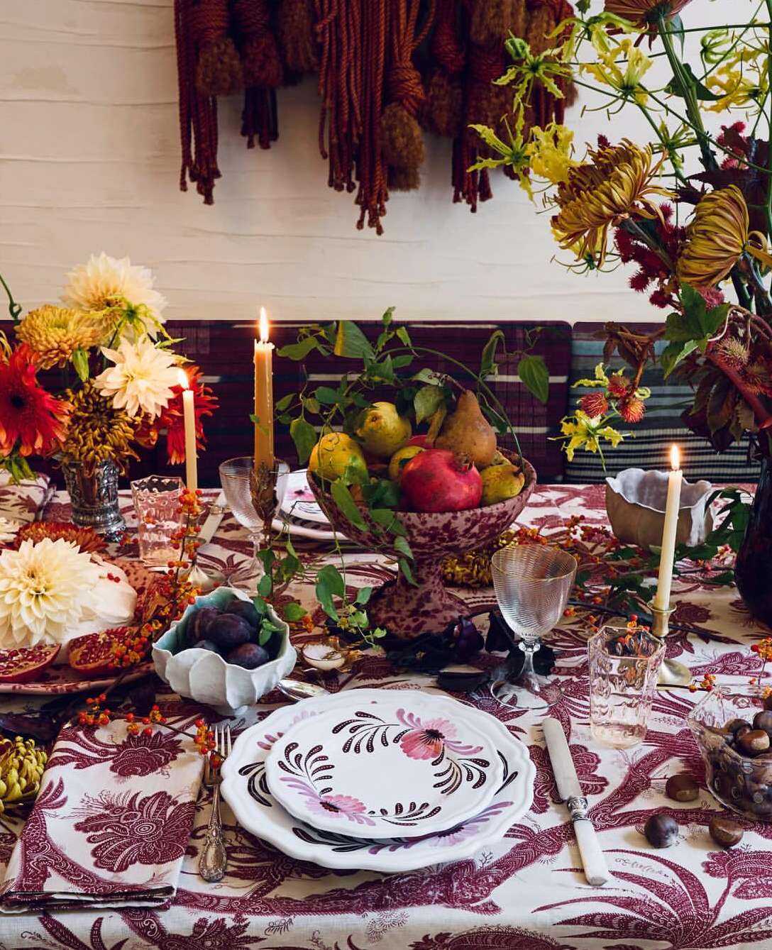 Table set for a feast with flowers, fruit, candles, plates and silverware on a floral tablecloth, creating a warm, inviting atmosphere.