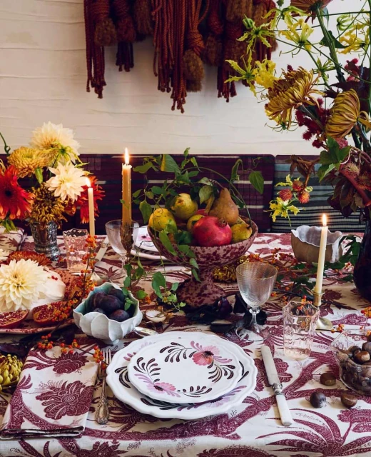 Table set for a feast with flowers, fruit, candles, plates and silverware on a floral tablecloth, creating a warm, inviting atmosphere.