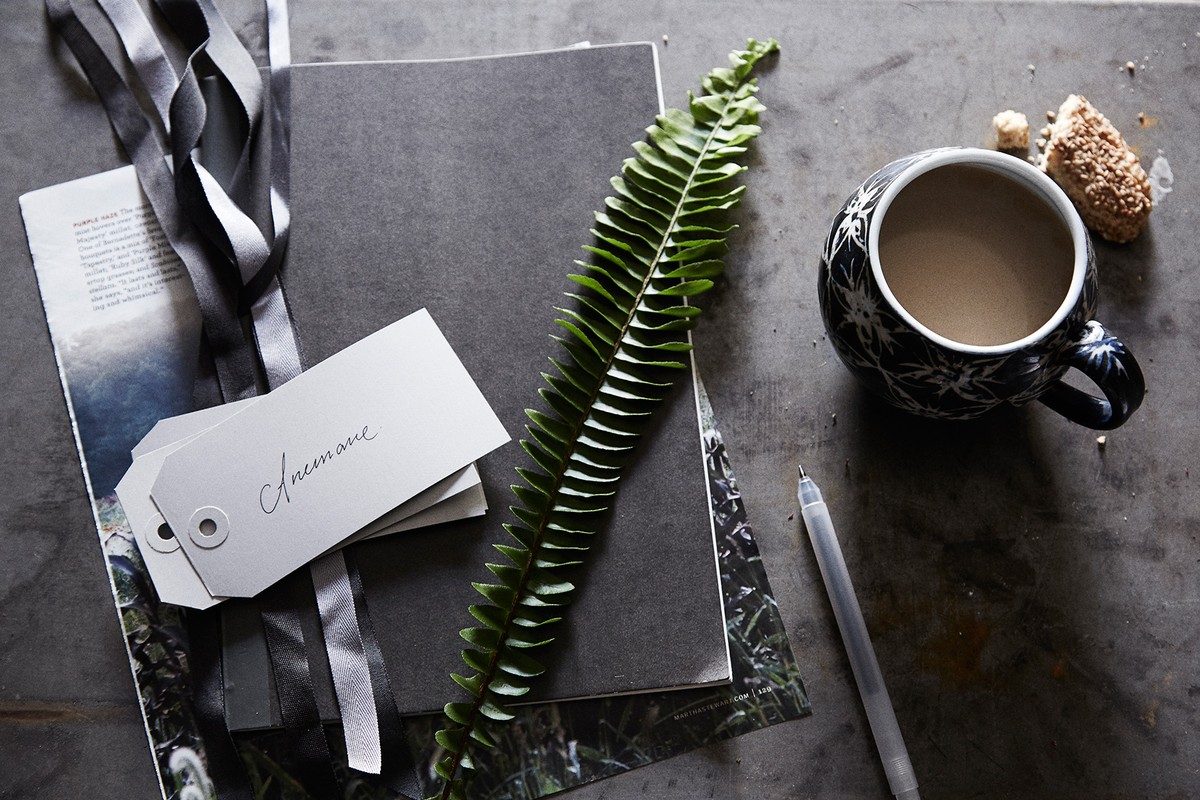 Still life featuring a coffee mug, fern, notebook, pen, and tags on a grey surface with a magazine, capturing a cozy, organized scene.