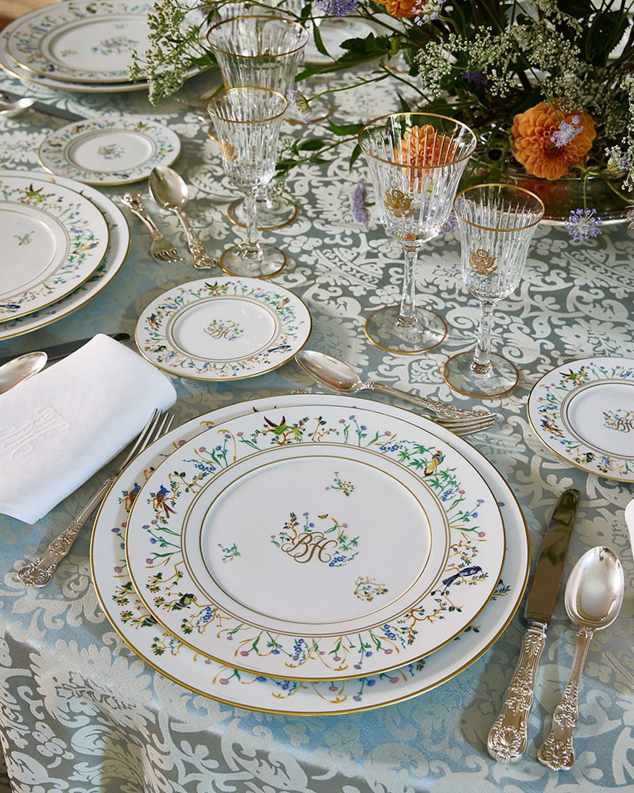 Ornate table setting with floral-rimmed china plates, crystal glasses, silver cutlery, and a white linen napkin on a patterned tablecloth.