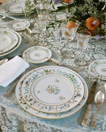 Ornate table setting with floral-rimmed china plates, crystal glasses, silver cutlery, and a white linen napkin on a patterned tablecloth.