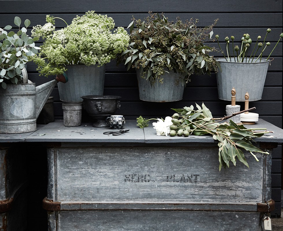 Rustic garden scene featuring galvanized buckets filled with various flowers and greenery on a vintage metal table against a dark wooden fence.