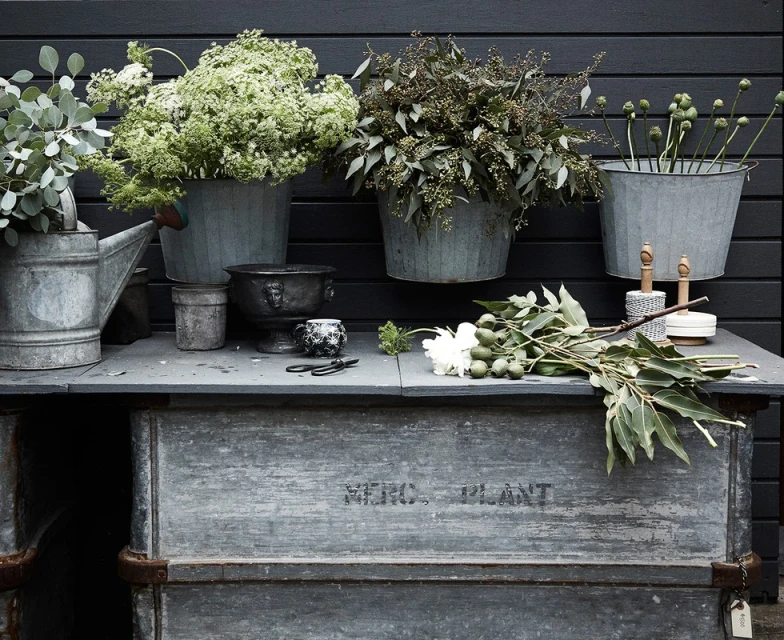 Rustic garden scene featuring galvanized buckets filled with various flowers and greenery on a vintage metal table against a dark wooden fence.