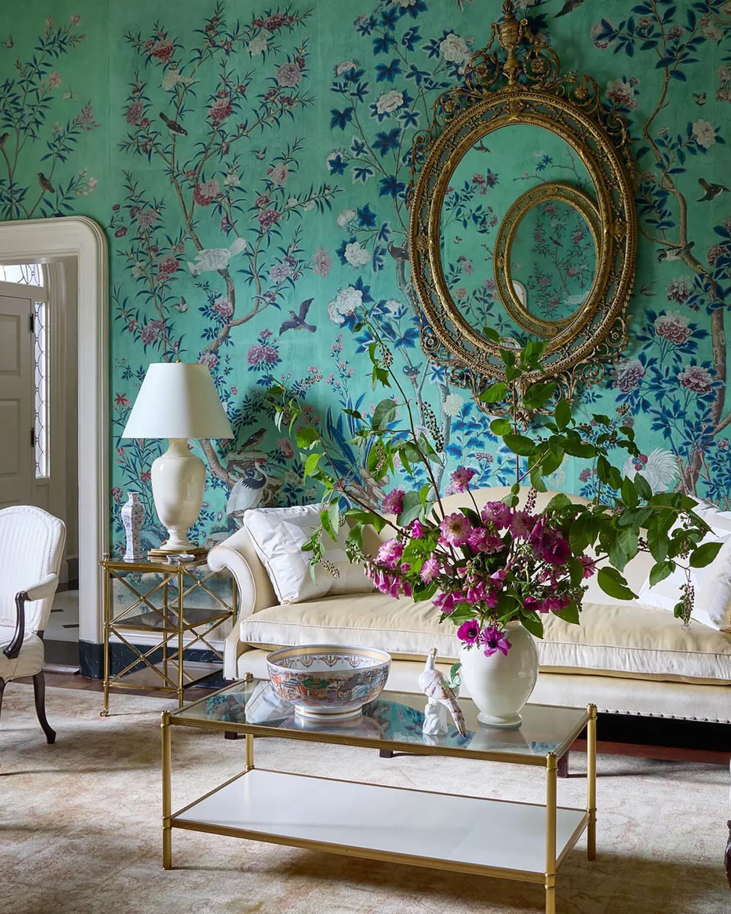Ornate living room interior featuring floral wallpaper, vintage furniture, a vase of pink flowers, and a decorative mirror.