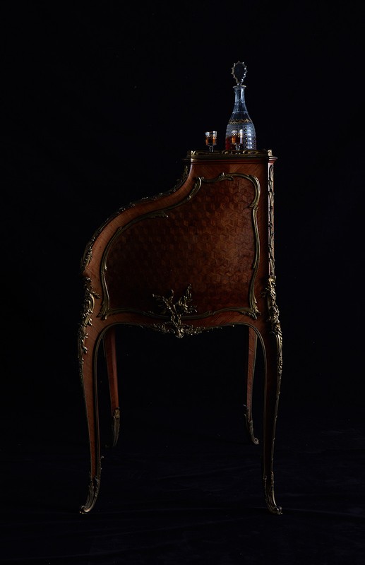 Ornate wooden writing desk with gilded trim and a crystal decanter, displayed against a stark black background, highlighting the desk's craftsmanship.