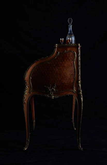 Ornate wooden writing desk with gilded trim and a crystal decanter, displayed against a stark black background, highlighting the desk's craftsmanship.