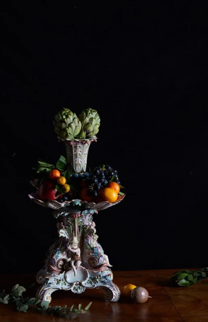 Ornate porcelain pedestal topped with vase holding artichokes, surrounded by mixed fruit, against a dark backdrop.