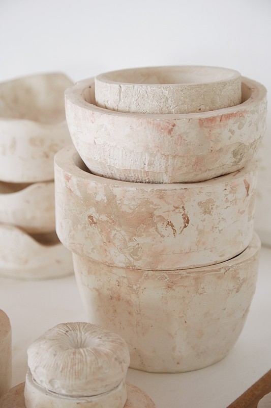Arrangement of handmade, beige-toned pottery bowls and covered containers on a white surface in a studio setting, with minimalist styling.