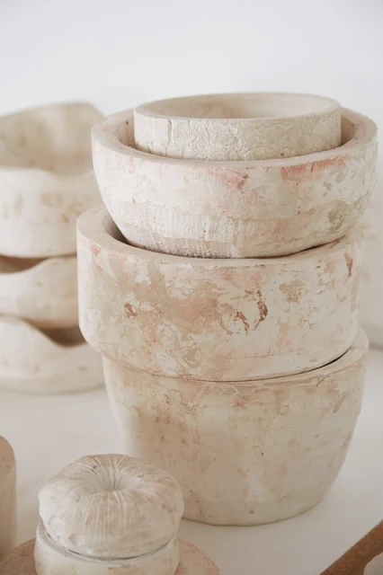 Arrangement of handmade, beige-toned pottery bowls and covered containers on a white surface in a studio setting, with minimalist styling.