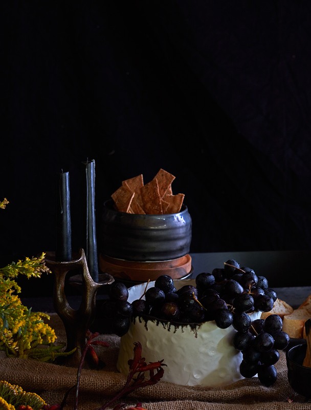 Still life featuring cheese with grapes, black candles in a holder, and crackers in a small bowl set against a dark background.