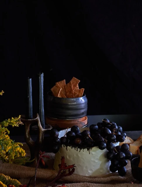 Still life featuring cheese with grapes, black candles in a holder, and crackers in a small bowl set against a dark background.