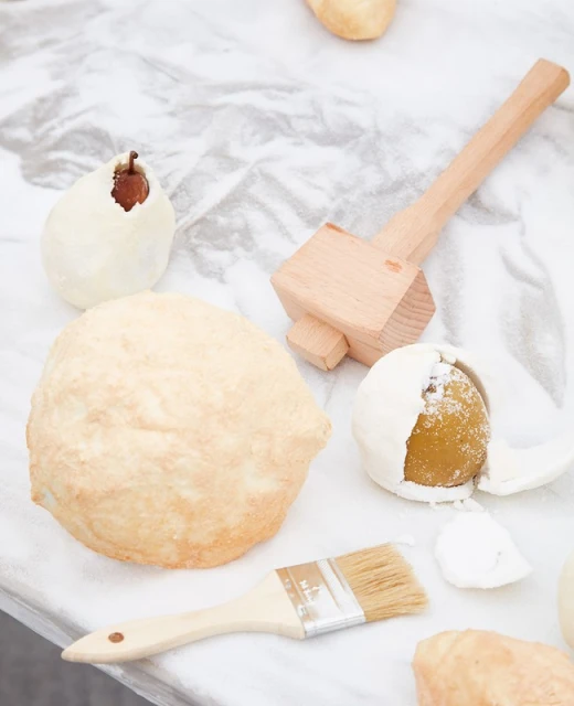 Overhead shot of food preparation scene featuring dough, wooden mallet, and pastry brush on a white marble surface, culinary setting.