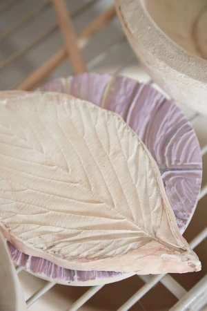 Close-up of carved leaf and textured bowl. Light beige and muted purple tones create a calm, artistic aesthetic in a studio setting.