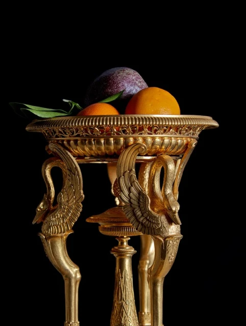 Golden footed bowl contains oranges, plum, and green leaves against a black background. Decorative gilded swan legs support the bowl.