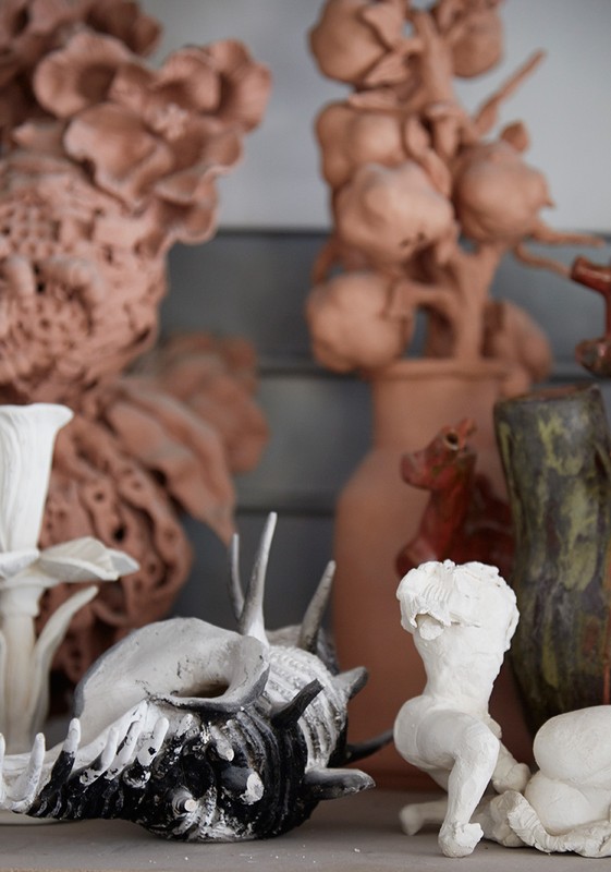 Collection of unpainted ceramic and clay art pieces including vases, figurative sculptures, and floral arrangements on a shelf.
