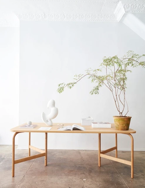 Minimalist interior features a light wood desk with a ceramic sculpture, open books, and a potted plant against a white wall.