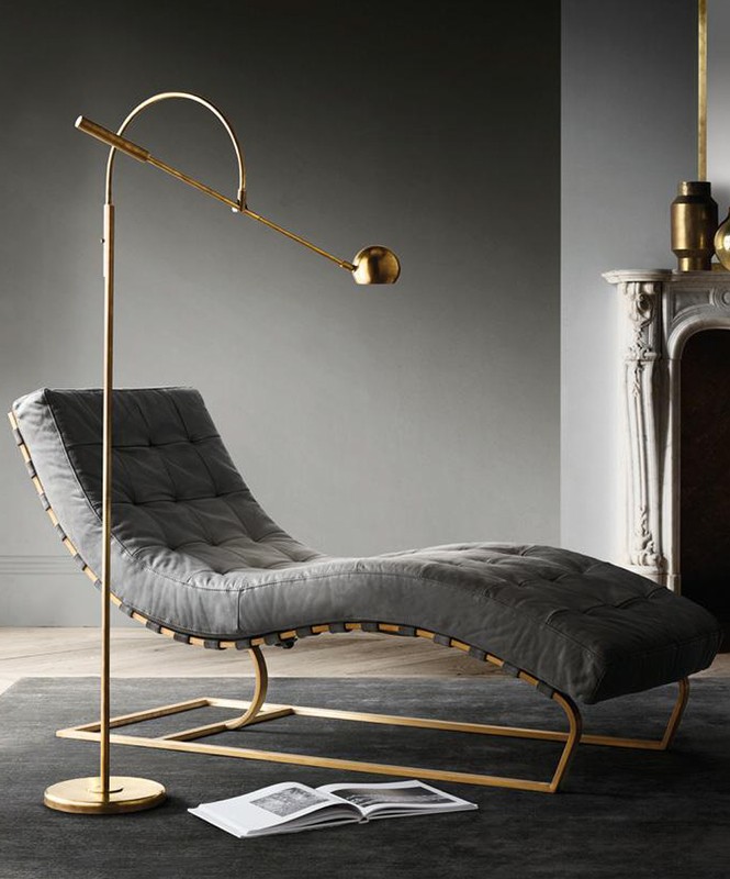 Gray tufted chaise lounge with gold frame and floor lamp near a fireplace, illuminated by natural light in an indoor setting.