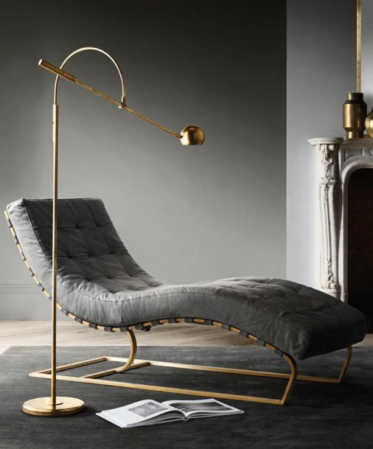 Gray tufted chaise lounge with gold frame and floor lamp near a fireplace, illuminated by natural light in an indoor setting.