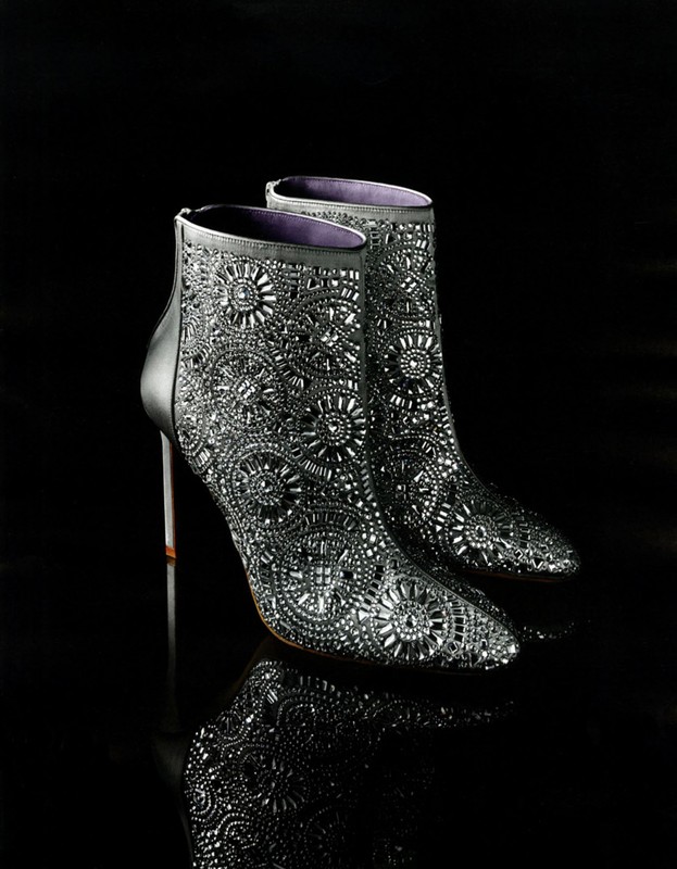 Elevated view of a pair of black, sparkling, ornate ankle boots with stiletto heels are displayed on a reflective black surface.